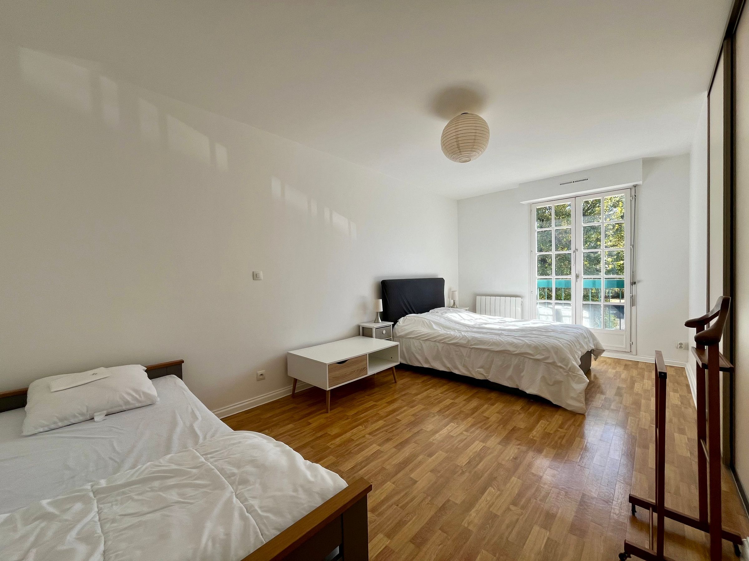 A bedroom with two beds, a bedside table, a radiator, and a window with a view of greenery.