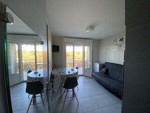 Una habitación con una mesa de comedor y sillas, un sofá y una televisión en la pared. Tiene un balcón con vistas al campo.