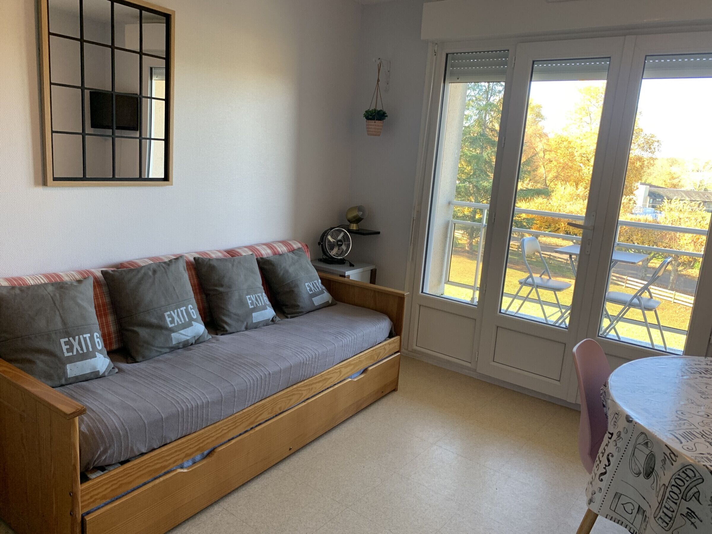 Ein Zimmer mit einer Schlafsofa aus Holz, einem kleinen Tisch mit karierter Tischdecke, einem Ventilator und Blick auf eine Außenpatio mit Stühlen und Tischen.