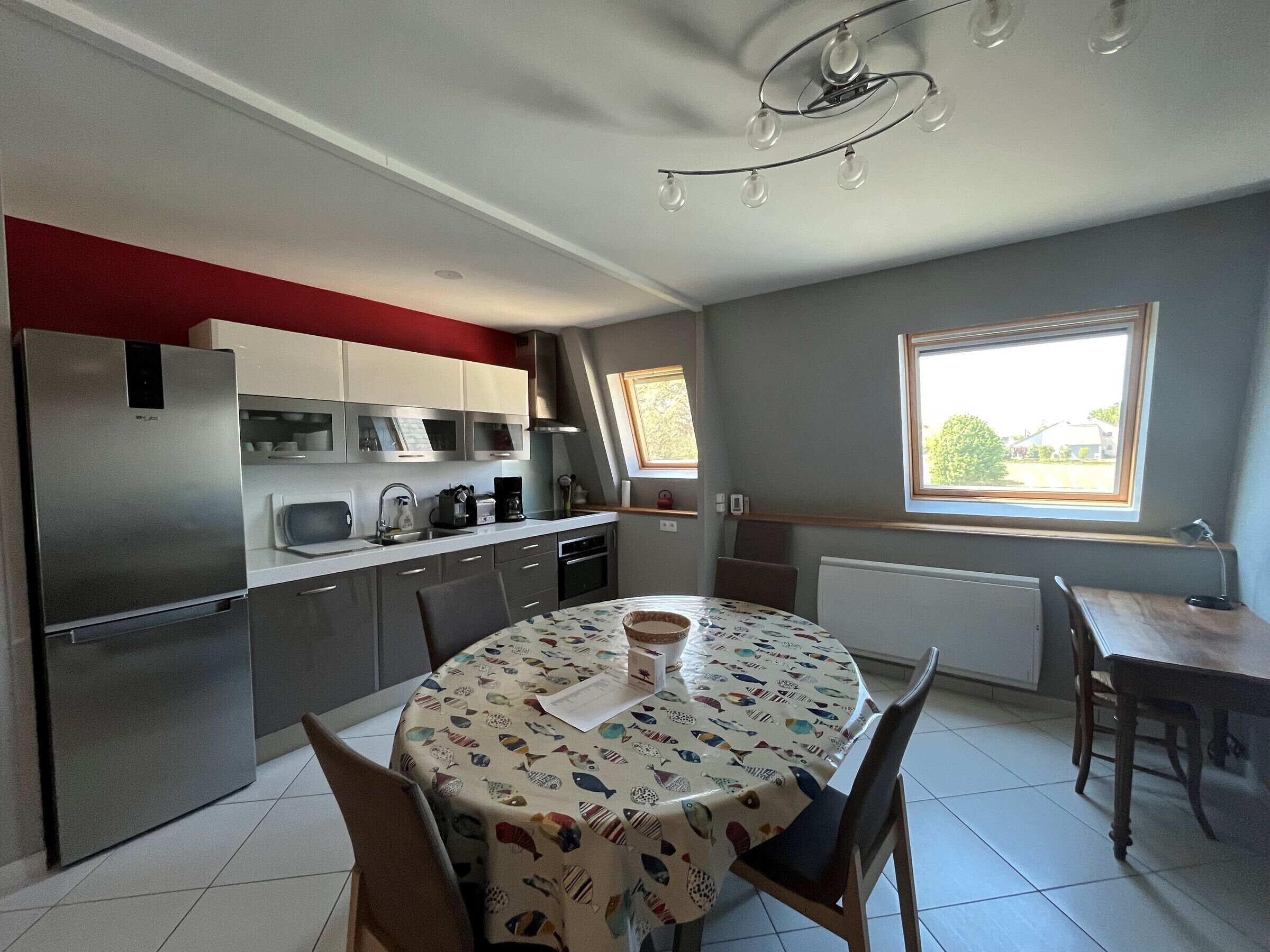 A compact kitchen with modern appliances, a dining table with a fish-patterned tablecloth, and a window providing natural light.