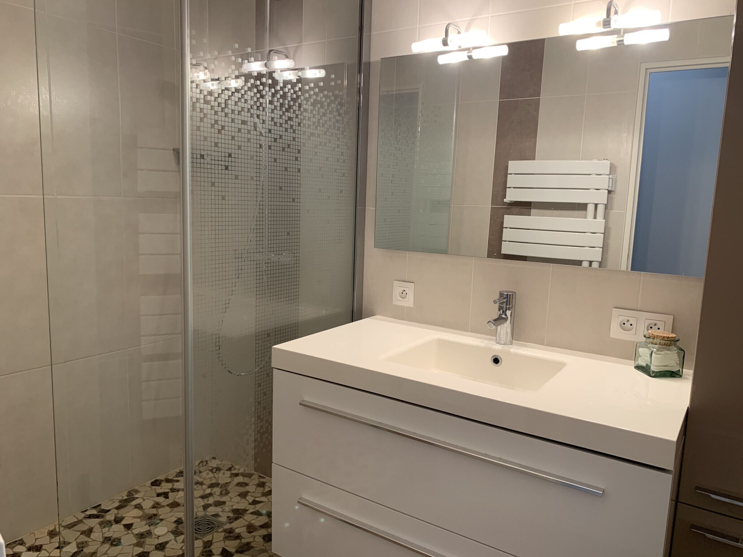 A modern bathroom with a glass shower enclosure, a white sink with a mirror above it, and a tiled floor.