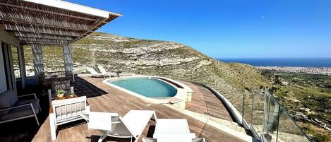 A pool area with lounge chairs and a pergola, overlooking a coastal landscape.