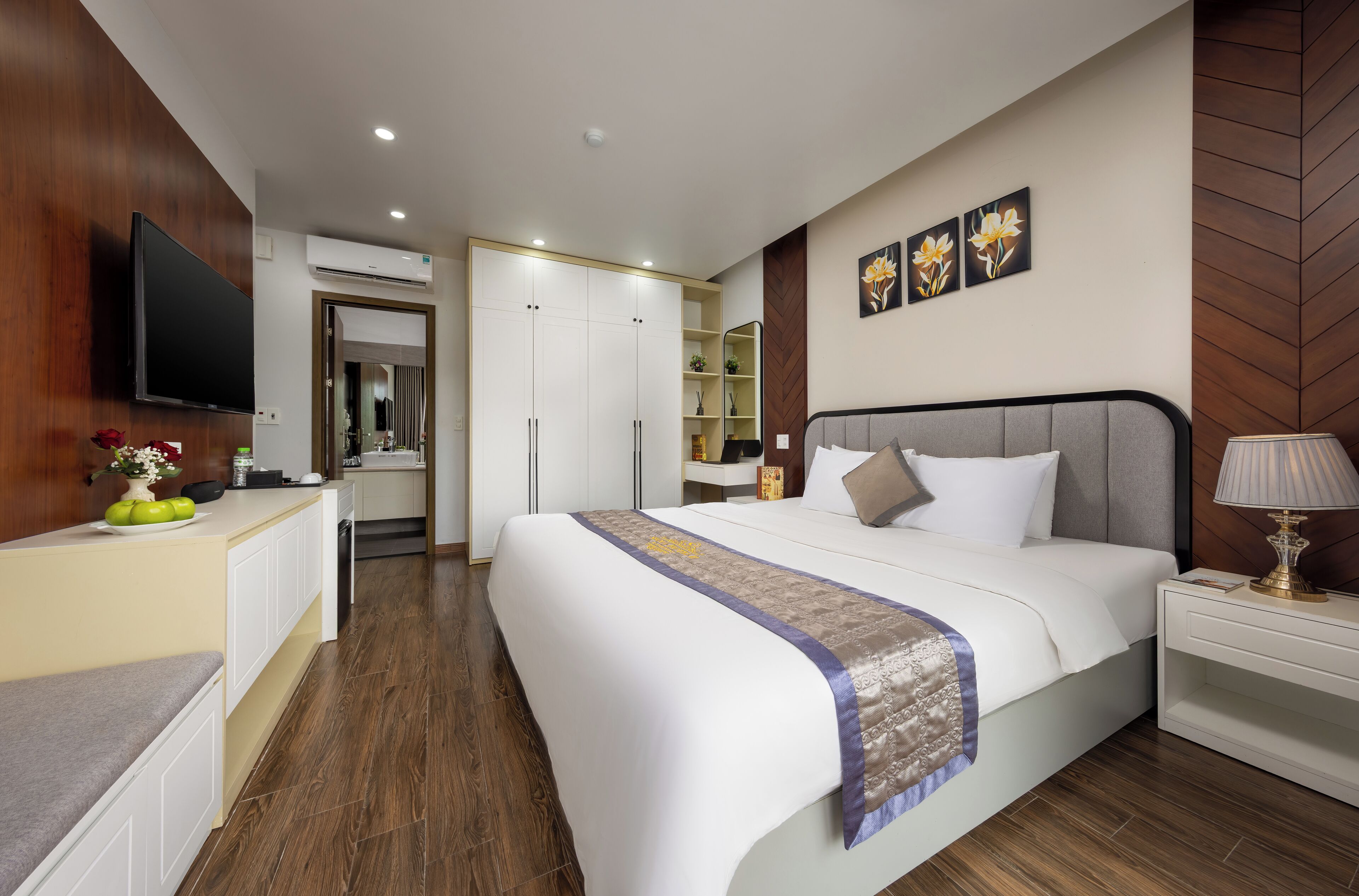 A modern hotel room with a large bed, a flat-screen TV mounted on the wall, a bedside table with a lamp, and a built-in wardrobe.