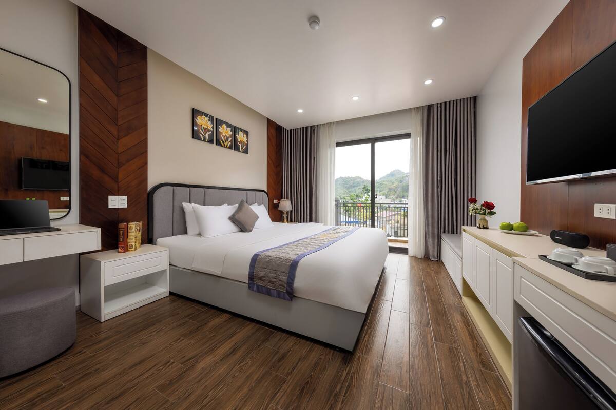 A modern hotel room with a large bed, a TV, a desk, and a balcony with a view of the mountains.