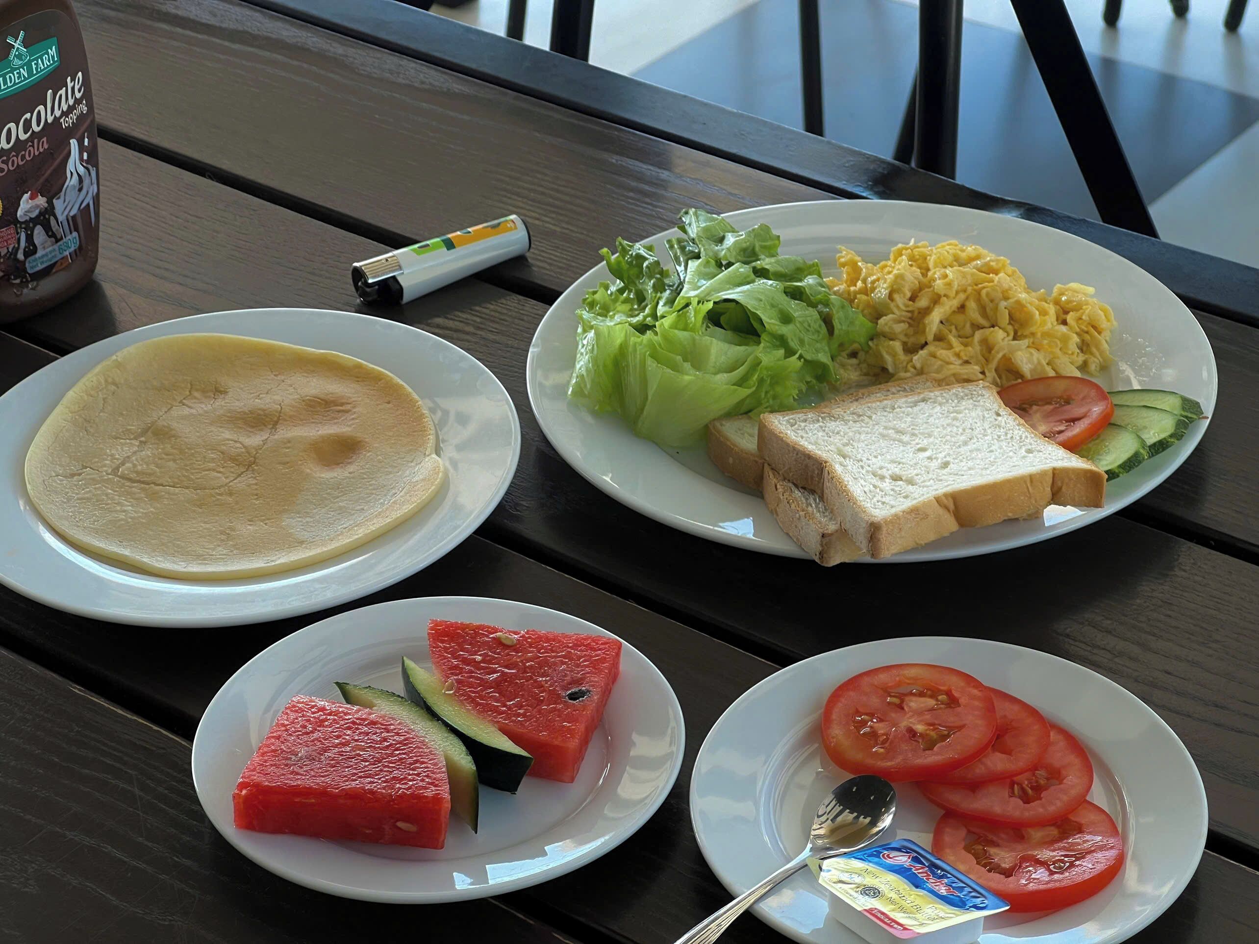 A table with plates of food including pancakes, scrambled eggs, toast, lettuce, cucumber, tomato, and watermelon.