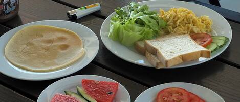 A table with plates of food including pancakes, scrambled eggs, toast, lettuce, cucumber, tomato, and watermelon.