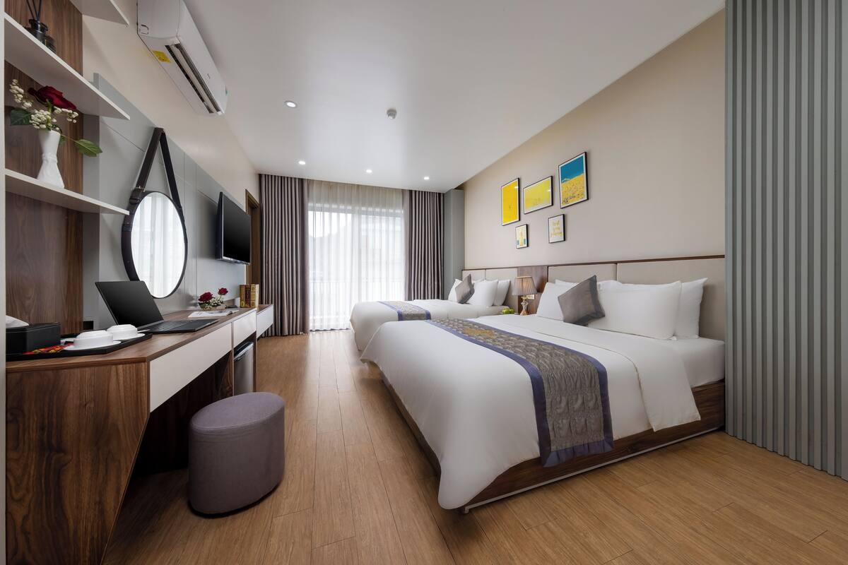 A modern hotel room with a wooden desk, a laptop, a flat-screen TV, a bed with white linens, and a large window with curtains.