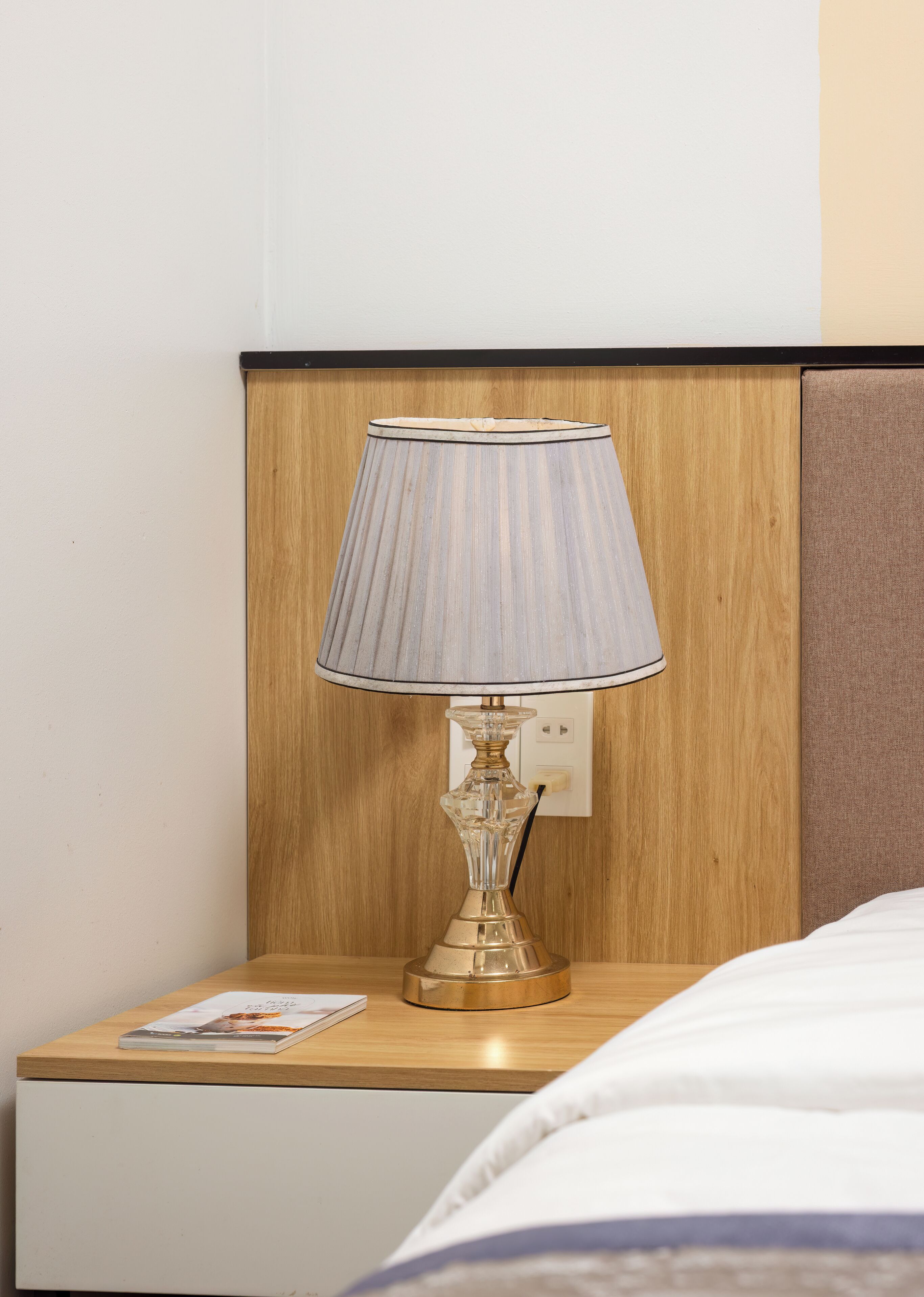 A bedside table with a lamp, a magazine, and a power outlet.