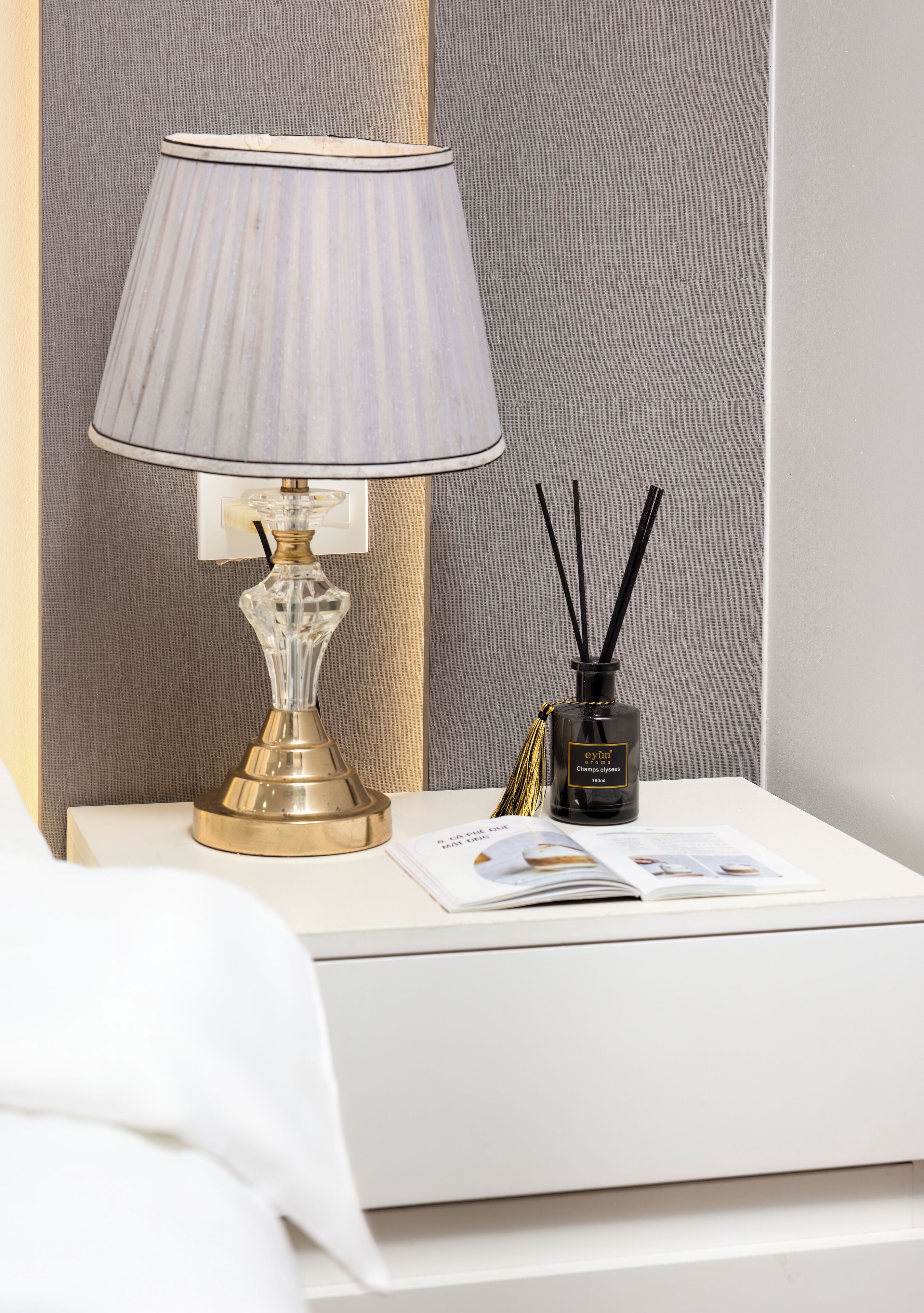 A bedside table with a lamp, a bottle of perfume, and a magazine.