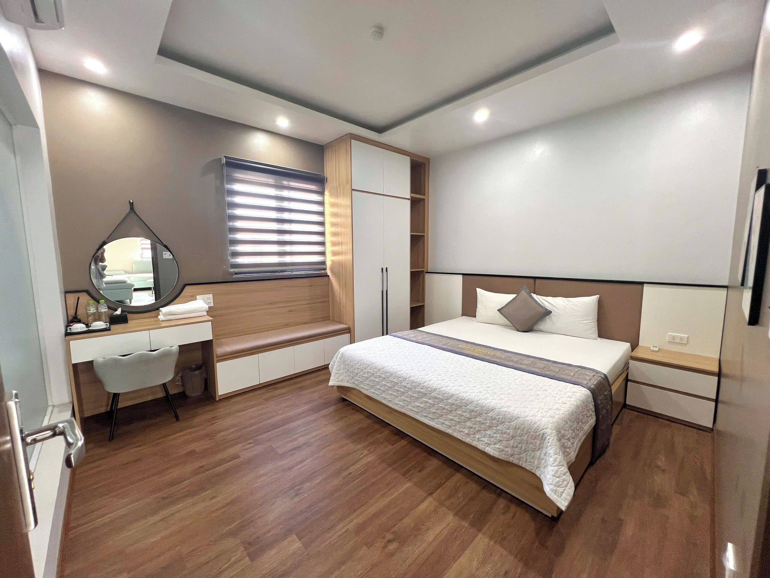A modern bedroom with a large bed, a bedside table, a wardrobe, a vanity with a mirror, and a bench.