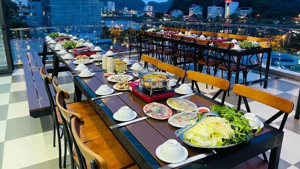 A rooftop dining area with multiple tables set for a meal, surrounded by chairs, and overlooking a cityscape with mountains in the background. - Cat Ba Seoul 2 Hotel & Apartment (Hai Phong)
