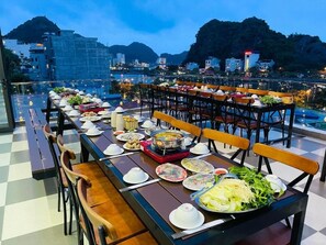 A rooftop dining area with multiple tables set for a meal, surrounded by chairs, and overlooking a cityscape with mountains in the background. - Cat Ba Seoul 2 Hotel & Apartment (Hai Phong)