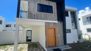 A modern two-story house with a wooden door, a balcony, and a small garden area. - Sky I Village Bavaro (Punta Cana)