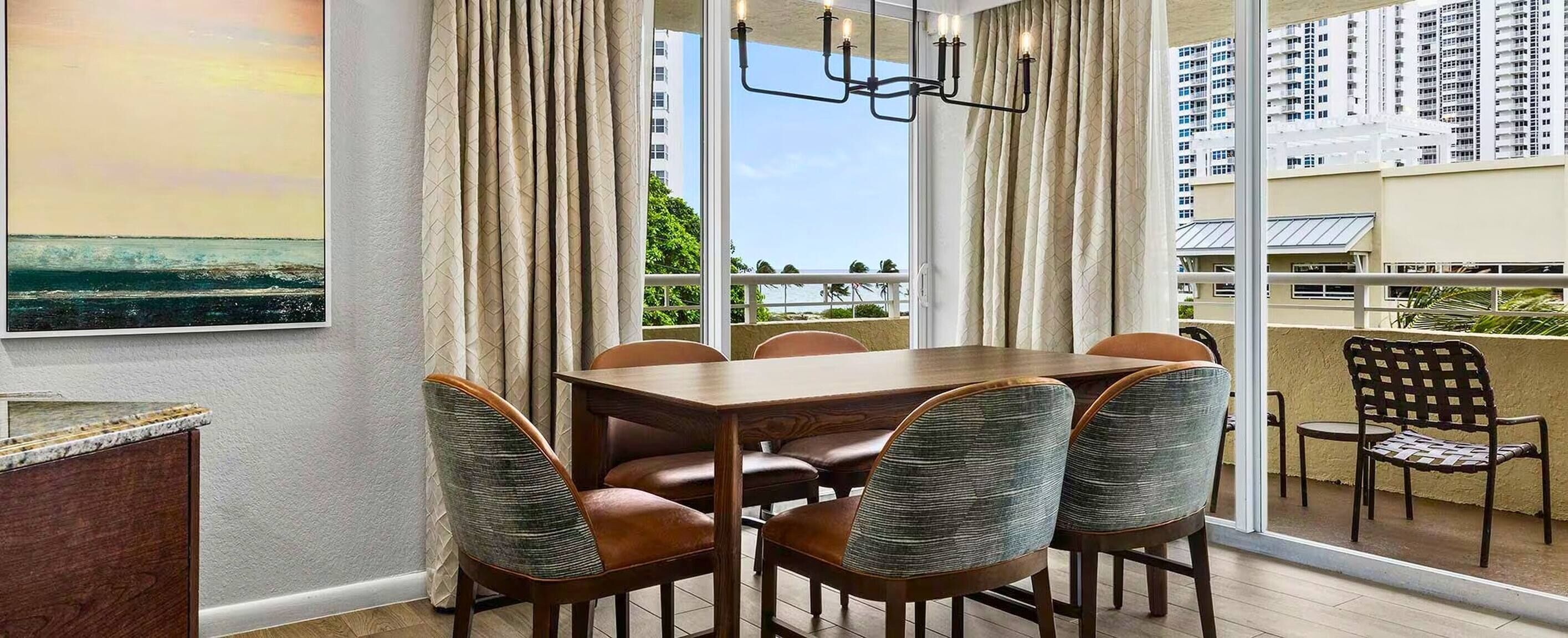 A dining area with a table and chairs, large windows with curtains, and a view of buildings and greenery.
