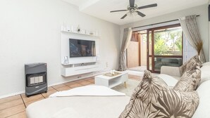 Un salón con televisor de pantalla plana, ventilador de techo, calefactor y sofá con cojines estampados.
