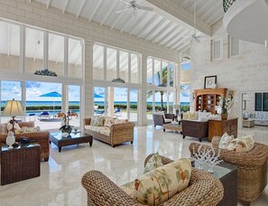 A spacious living area with wicker furniture, large windows offering ocean views, and a ceiling fan. - Ocean Mist Villa - by Zenbreak (Long Bay)