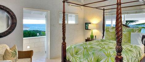 Una cama con dosel, un estampado de hojas tropicales, una silla de mimbre y vistas al océano.