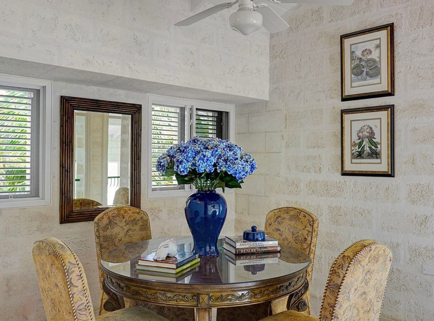 Un comedor con una mesa redonda de vidrio, cuatro sillas tapizadas, un jarrón azul con flores e ilustraciones botánicas enmarcadas en la pared.