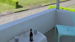 A balcony with a view of a green field and a modern building in the distance. There is a small table with a bottle and cans on it, and a light blue chair. - In Espinal Casaquinta (Espinal)