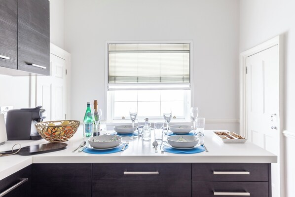 A modern kitchen with white cabinets, a central island with blue placemats, and a window with blinds. - SOHO - Spacious 5 bed, 2bath Apartment-SOHO (London)