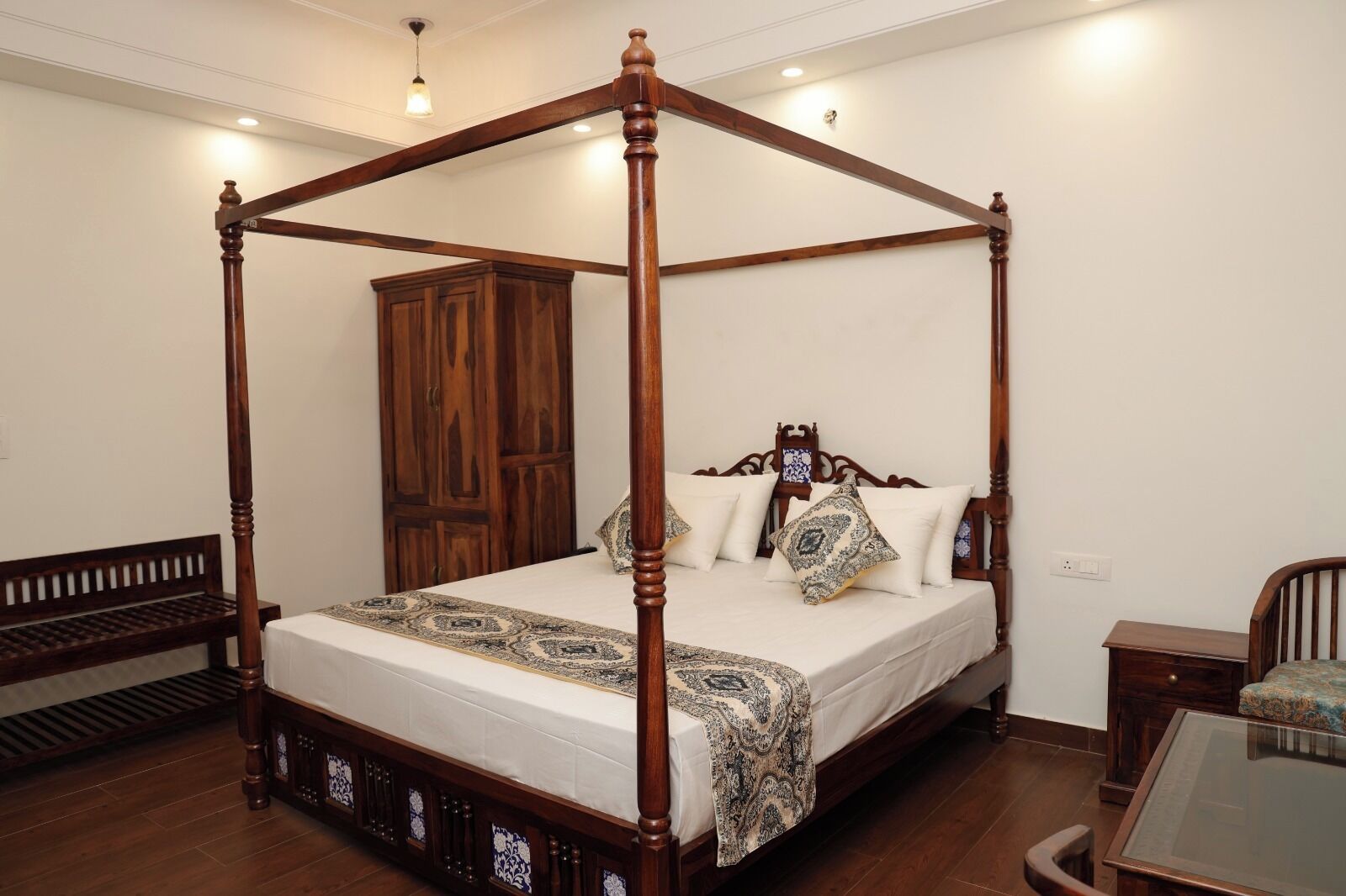 Una cama con dosel y cuatro columnas, con estructura y cabecera de madera, un armario grande, un banco y una mesa con superficie de vidrio.