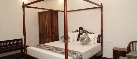 Una cama con dosel y cuatro columnas, con estructura y cabecera de madera, un armario grande, un banco y una mesa con superficie de vidrio.