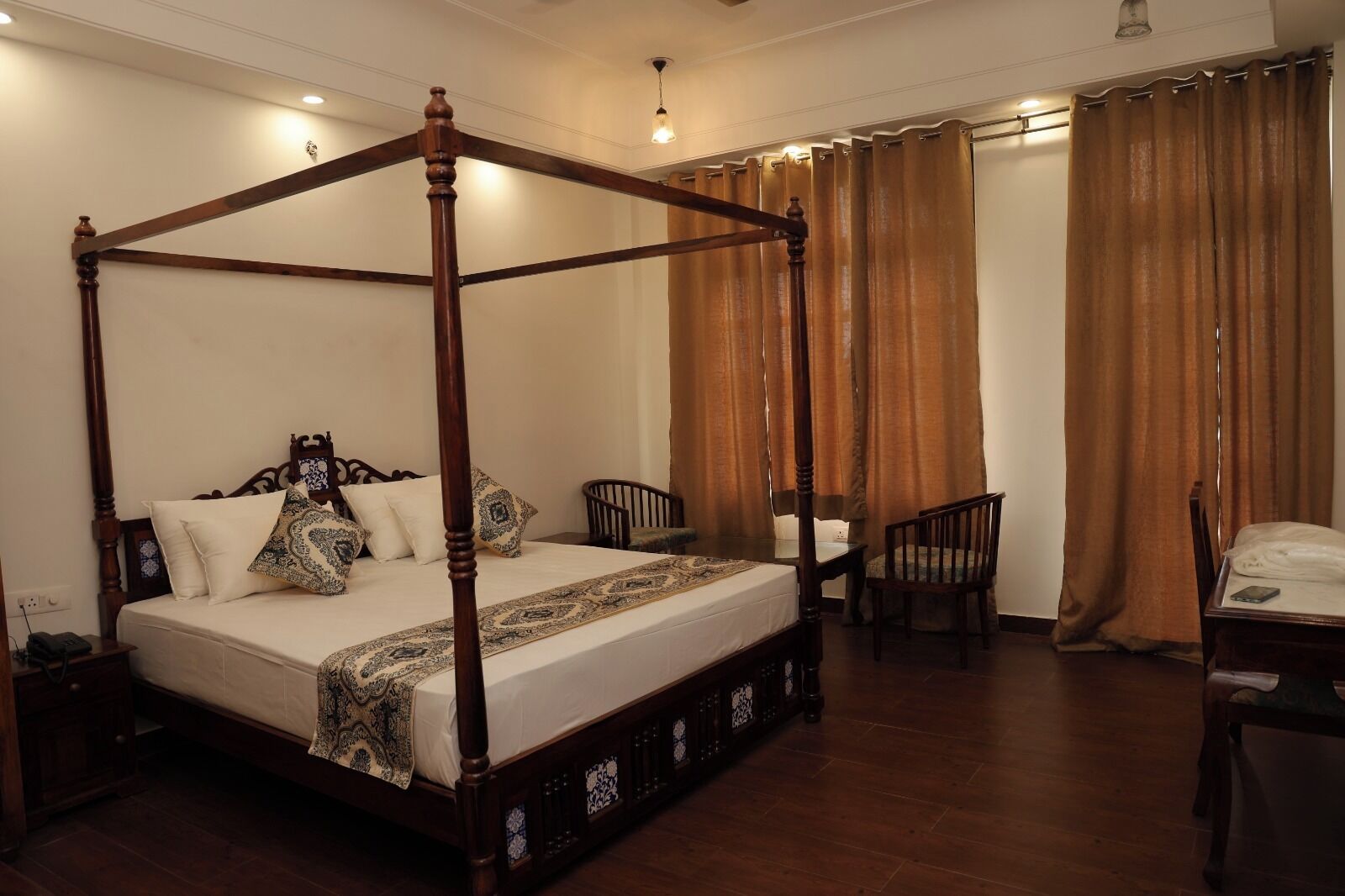 A four-poster bed with a wooden frame and a matching headboard, a bedside table, a chair, and a small table with a chair.