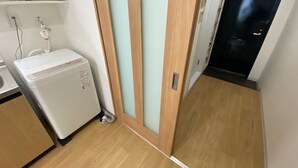 A compact kitchen with a refrigerator, a sink, and a cabinet. A wooden door with a frosted glass panel leads to a dark room. - W110 - Quaint Sakurashinmachi - Newly Renovated Unit with Two Beds  (Setagaya City)