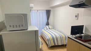 A compact hotel room with a kitchenette, a bed with striped bedding, a desk with a computer, and a window with curtains. - W110 - Quaint Sakurashinmachi - Newly Renovated Unit with Two Beds  (Setagaya City)