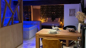 Een moderne slaapkamer met een groot bed, een kleine badkamer met een badkuip met blauw licht, en een eethoek met een houten tafel gedekt voor twee personen.