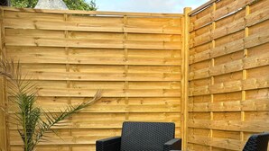 Een terras buiten met rieten meubels, waaronder een bank en twee fauteuils, en een kleine, zwarte tafel.