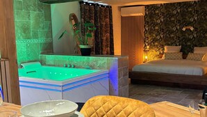 Een moderne hotelkamer met een wellnessruimte, een bed met veel kussens en een eethoek met een gedekte tafel voor twee.