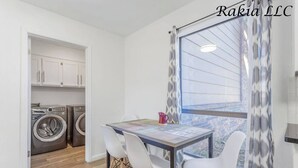 Un espace de vie compact comprenant un coin repas, un lave-linge et une vue sur un immeuble en briques, visible par une fenêtre.