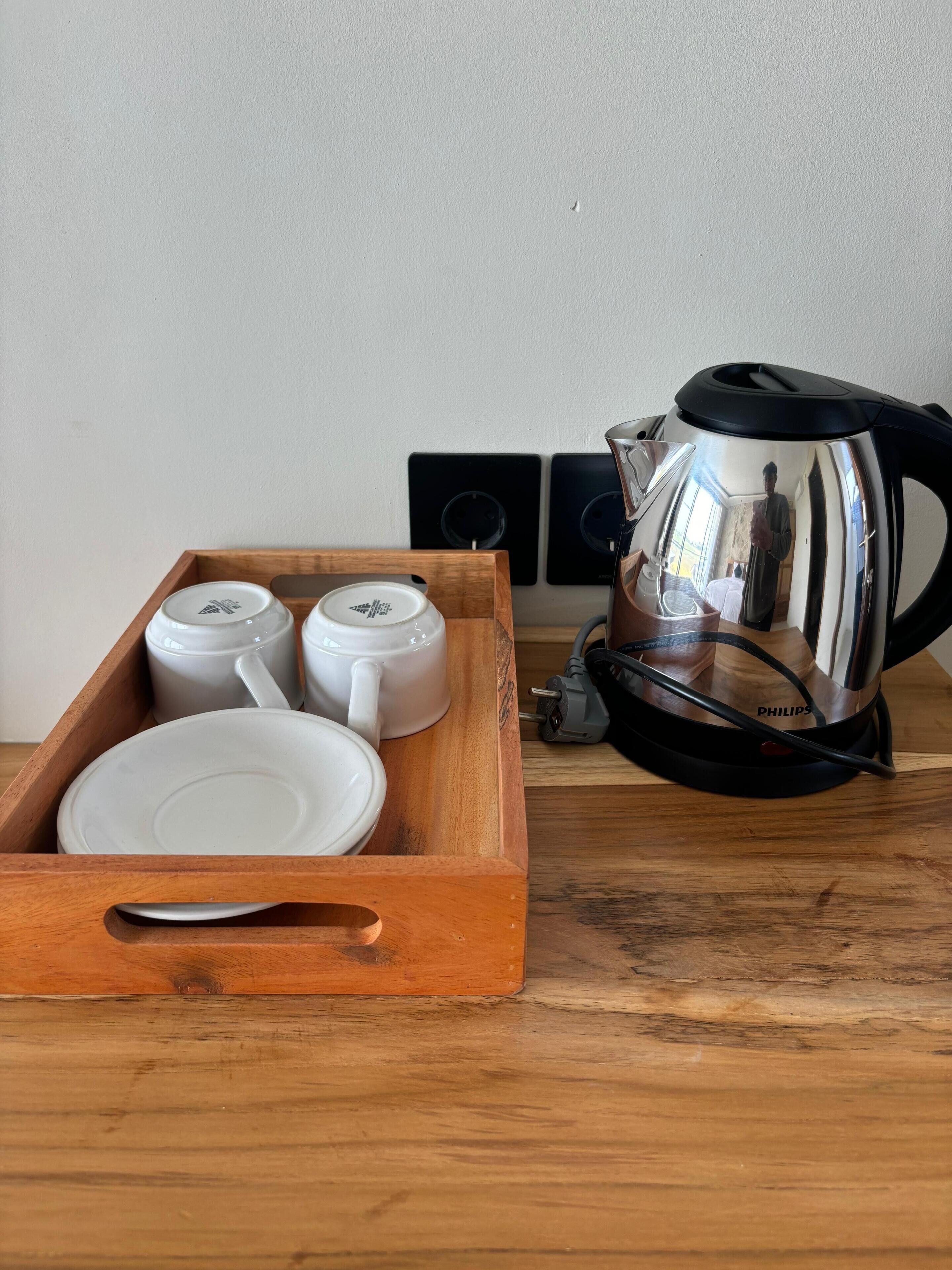 A wooden tray with white cups and a plate, a black and silver coffee maker, and a power strip with a plug on a wooden table.