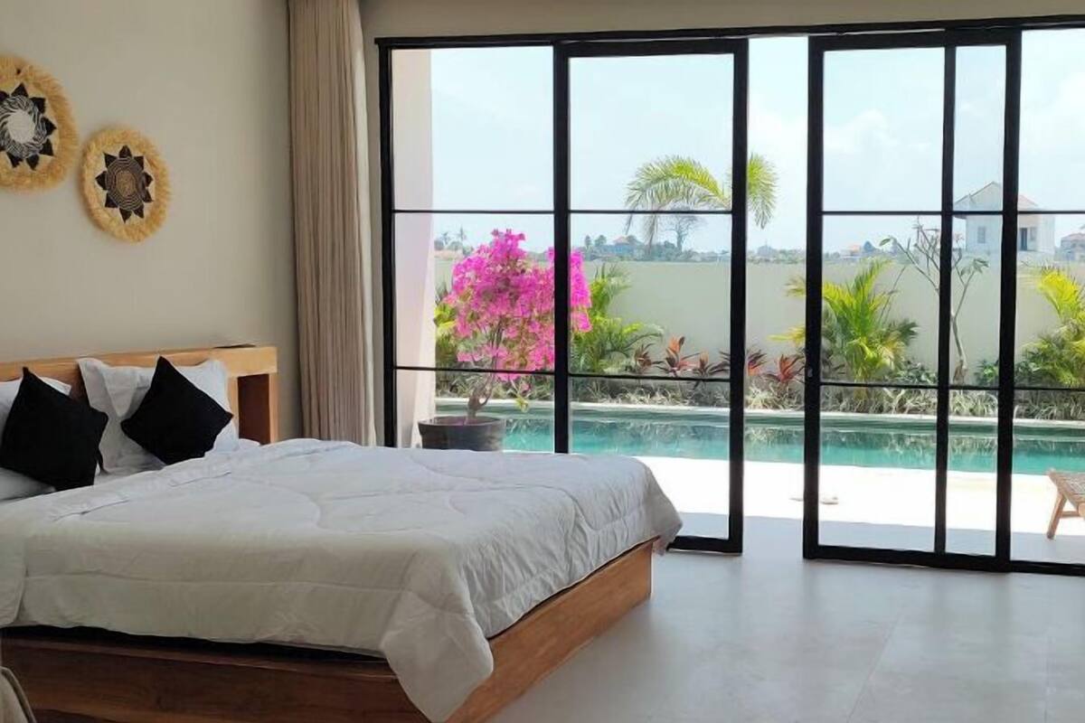 Ein Schlafzimmer mit einem großen Bett, Blick auf einen Swimmingpool und einem Balkon mit Pflanzen.