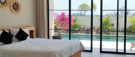 A bedroom with a large bed, a view of a swimming pool, and a balcony with plants.