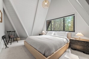 Een slaapkamer met een schuine kap, een groot raam, een bed met houten frame, twee nachtkastjes met lampen, en een stoel met een mand.
