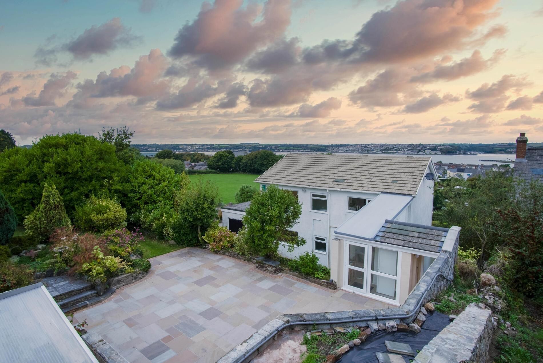 Ein modernes, zweistöckiges Haus mit einer großen Terrasse, umgeben von üppiger Vegetation und einem malerischen Blick auf das Meer und eine ferne Stadt.