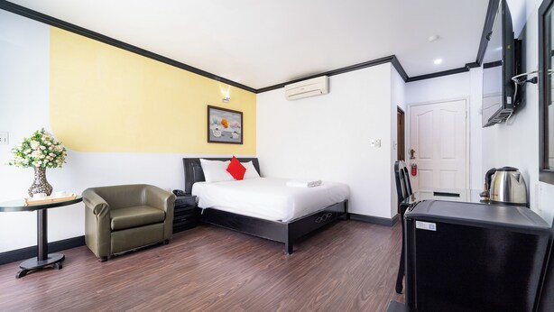 A compact hotel room with a bed, a small armchair, a desk with a kettle, and a TV mounted on the wall.