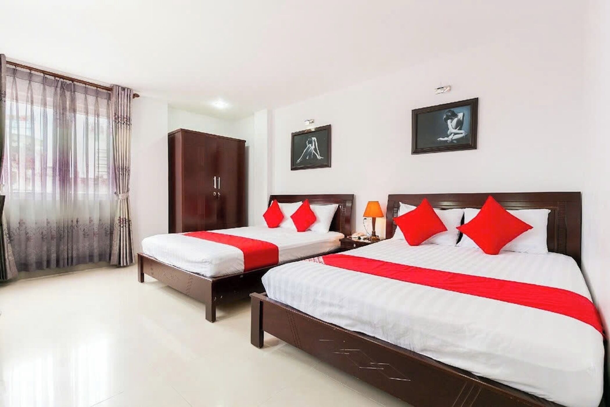 Quarto de hotel com duas camas, cada uma com almofadas vermelhas e um edredom branco. Há duas obras de arte emolduradas na parede, um guarda-roupa e uma janela com cortinas.