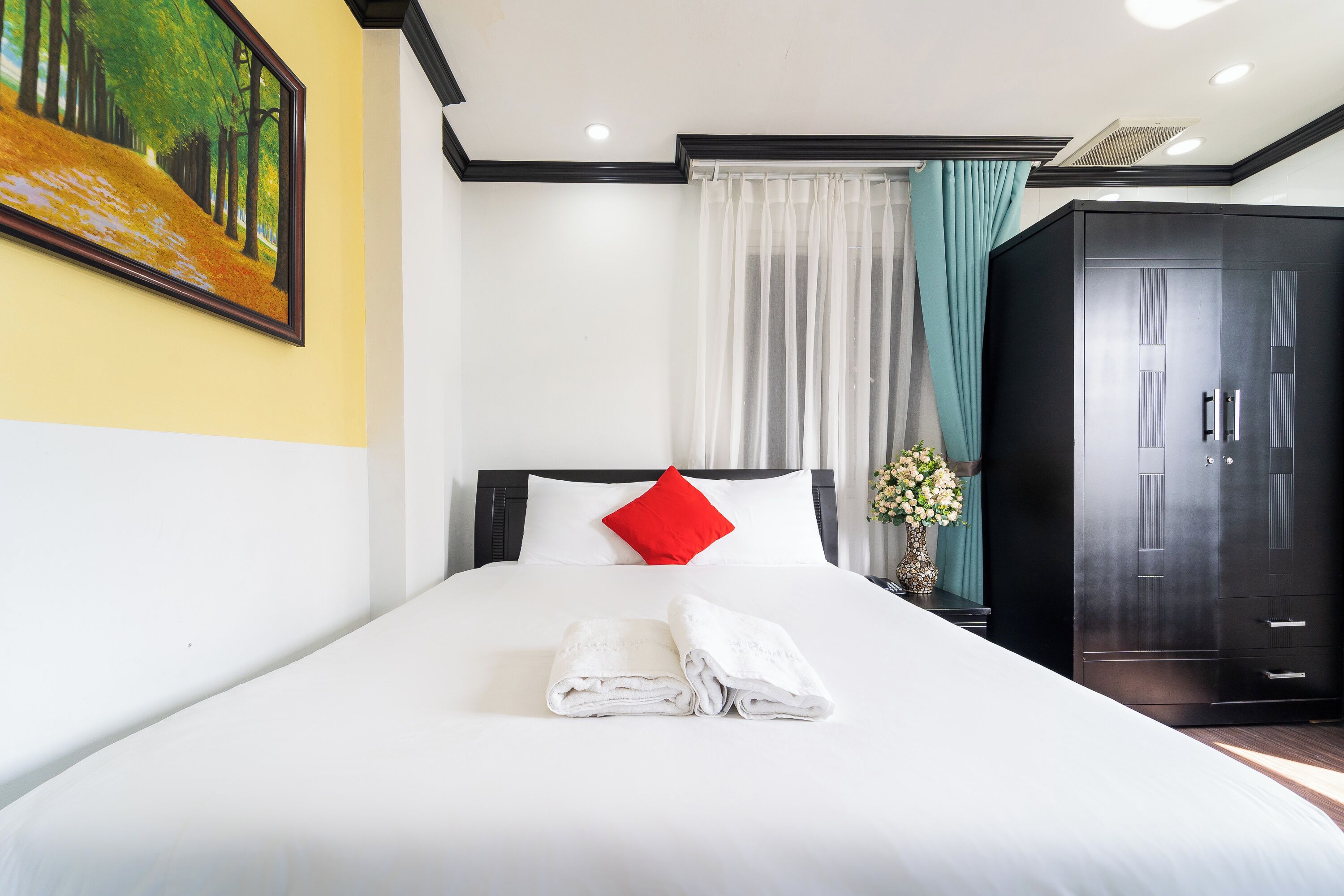 A hotel room with a neatly made bed, a red pillow, a large wardrobe, and a painting on the wall.