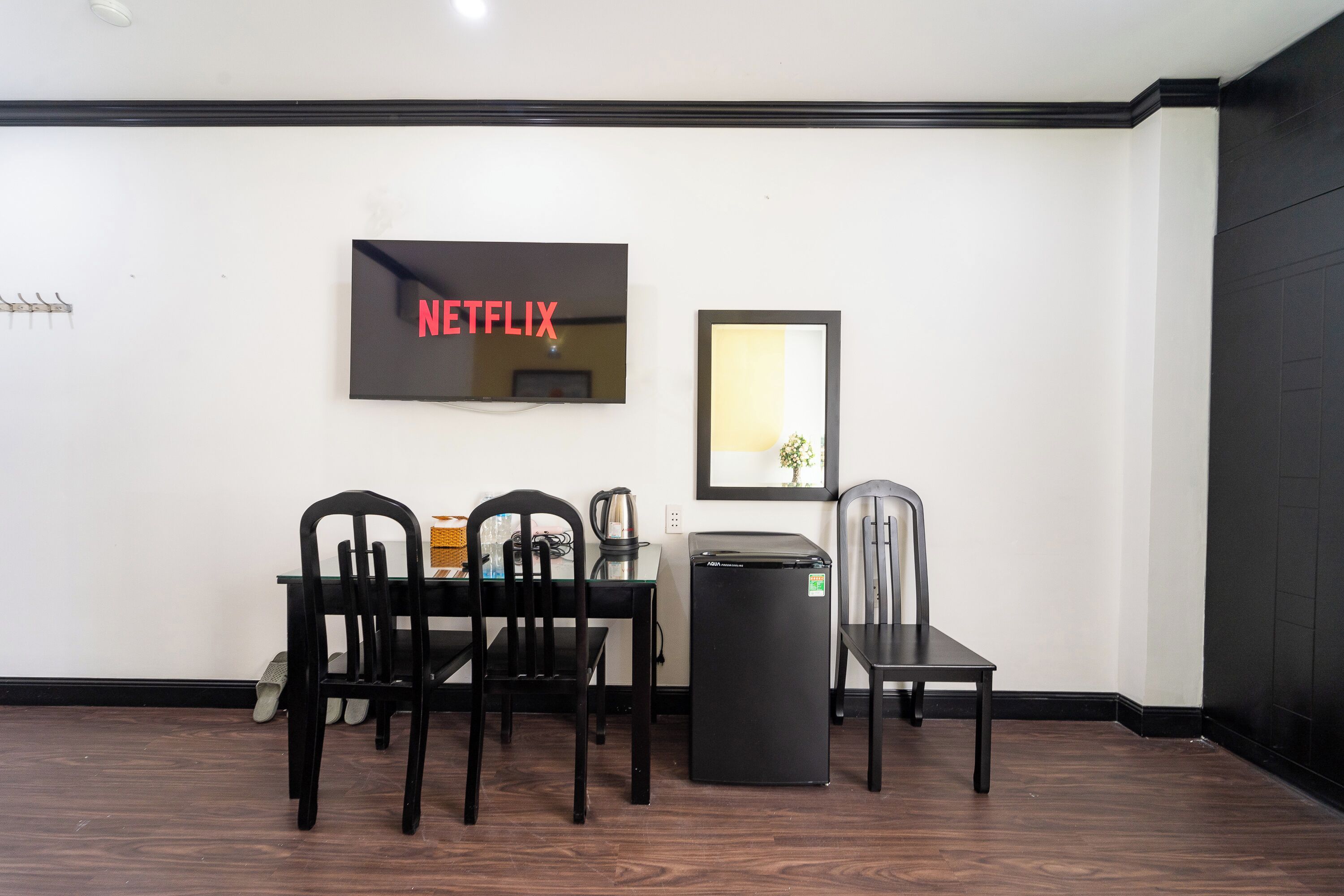 A room with a dining table, chairs, a refrigerator, a television mounted on the wall, and a framed picture.