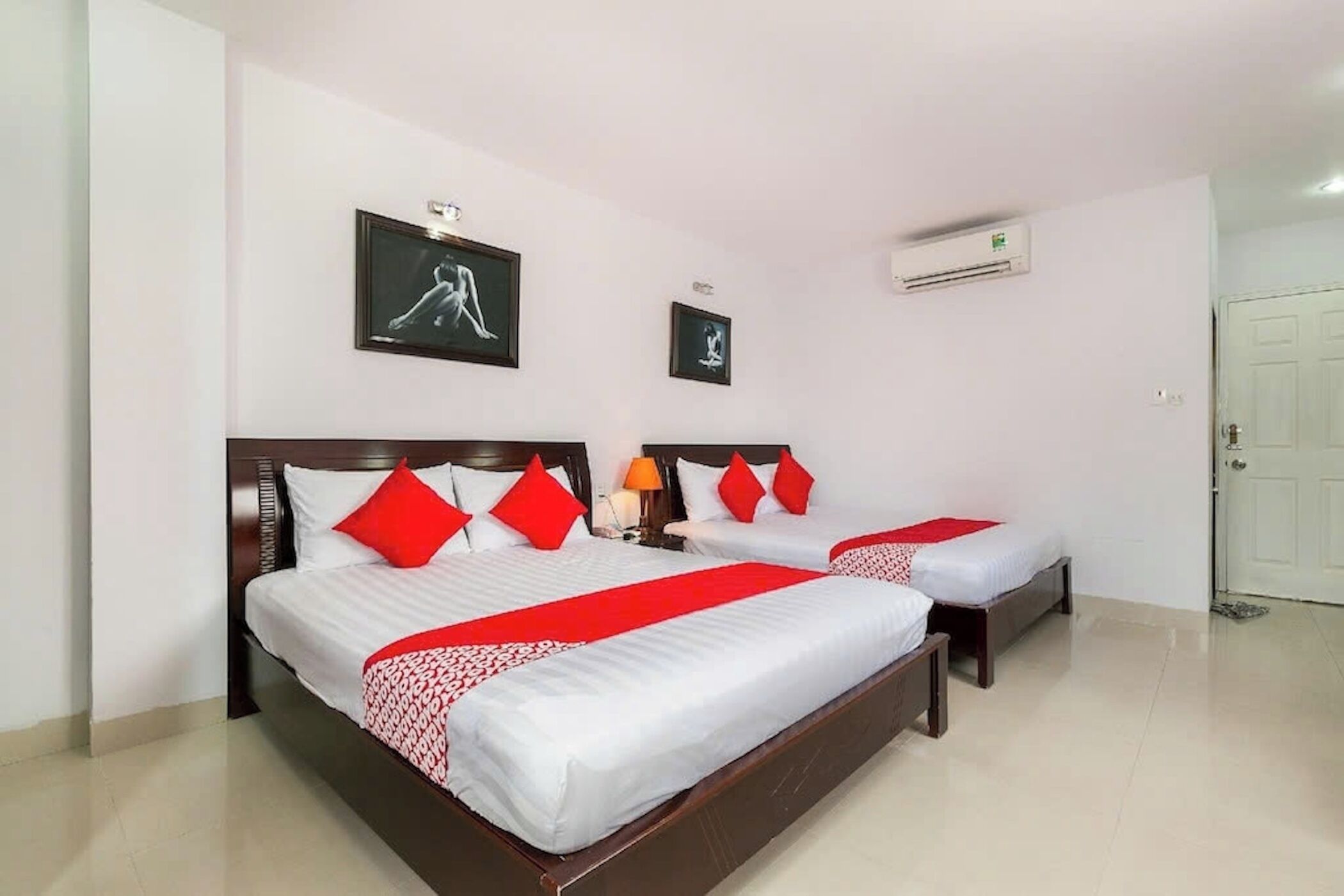 Habitación de hotel con dos camas, cada una con ropa blanca y detalles rojos, cabeceros de madera y aire acondicionado instalado en la pared.