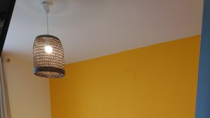 Une chambre avec un lit jaune, deux oreillers, une serviette blanche, un luminaire en osier et une petite table de chevet.