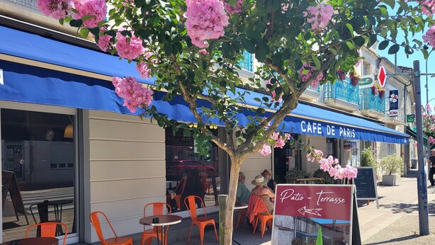 Un café en plein air avec des chaises et des tables orange, un auvent bleu et une enseigne indiquant « Patio – terrasse ».