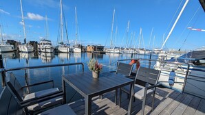 A marina with multiple sailboats docked. There is a wooden deck with a table and chairs, and a potted plant on the table. - Houseboat Fjord Stella with bio sauna and roof terrace in Wendtorf/ Kiel Fjord (Wendtorf)