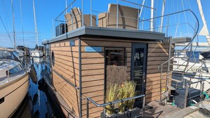 A small wooden structure with a glass door, named 'Fjord Stella', located at a marina with boats docked nearby. - Houseboat Fjord Stella with bio sauna and roof terrace in Wendtorf/ Kiel Fjord (Wendtorf)