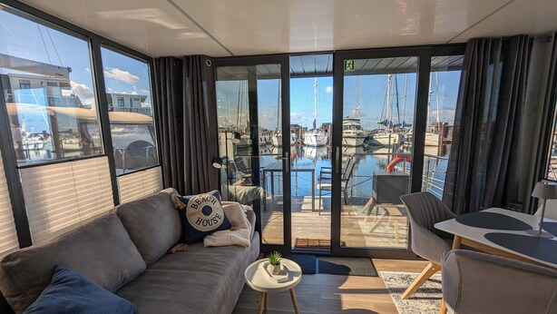 A modern living room with a sofa, a small table, and a view of a marina with boats. - Houseboat Fjord Stella with bio sauna and roof terrace in Wendtorf/ Kiel Fjord (Wendtorf)