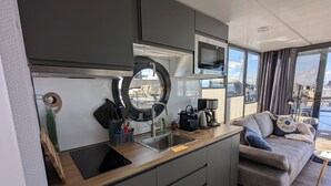 A compact kitchen with a sink, a coffee maker, and a microwave. There is a window with a view of boats and a sofa with a 'Beach House' pillow. - Houseboat Fjord Stella with bio sauna and roof terrace in Wendtorf/ Kiel Fjord (Wendtorf)