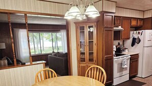 Una cocina compacta con armarios de madera, una estufa y un refrigerador. Hay una mesa de comedor con sillas y un ventanal con vistas a un espacio exterior arbolado.